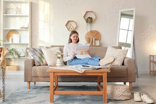 Beautiful young woman reading newspaper on sofa in living room