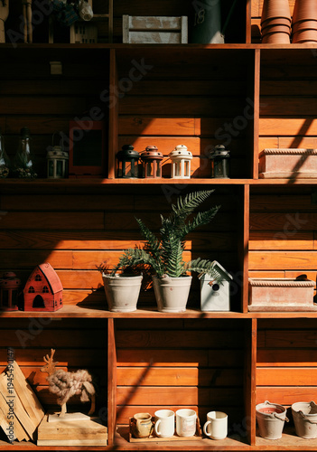 Wooden interior shelf decorated with plants, lanterns, pottery and small birdhouses in warm sunlight. Rustic home decoration concept with natural materials and cozy atmosphere.