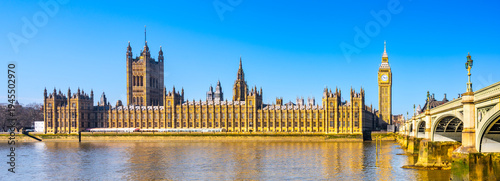 The Houses of Parliament and Big Ben along the River Thames. The sky is clear. People visit the area to admire this famous landmark.
