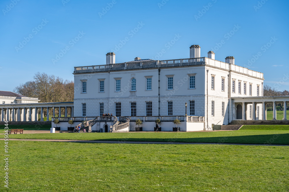 Fototapeta premium People explore Greenwich Queens House in London during a sunny day. The white building stands surrounded by green lawns and trees, showing the location's historical significance.