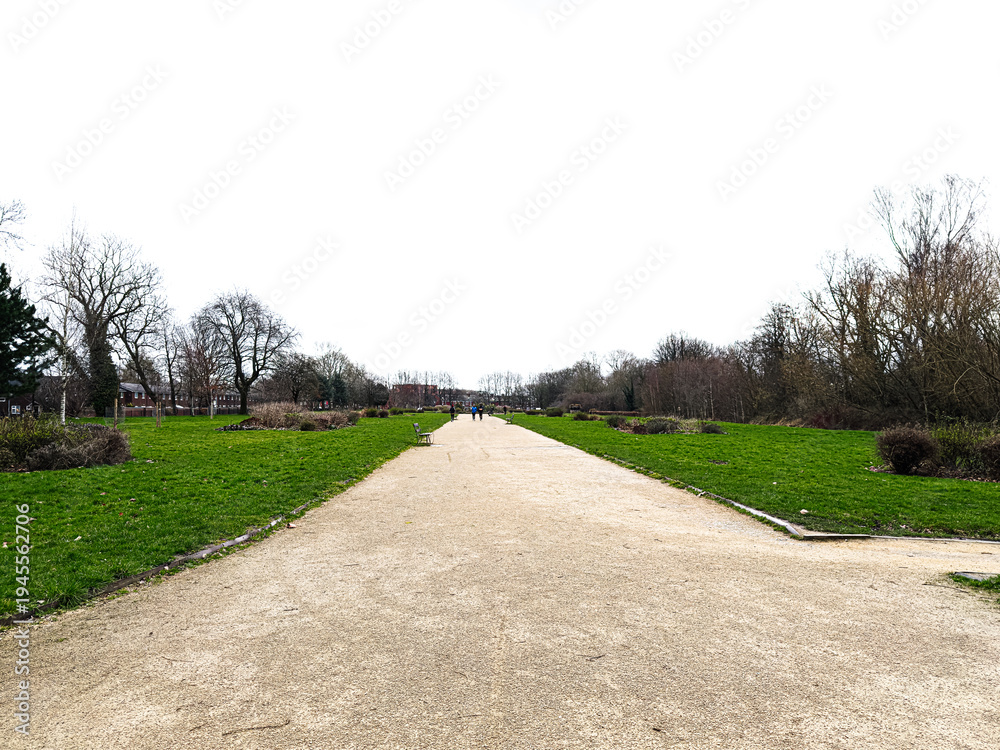 Obraz premium Wide gravel path through Alexandra Park in Manchester UK with benches, trees and distant walkers