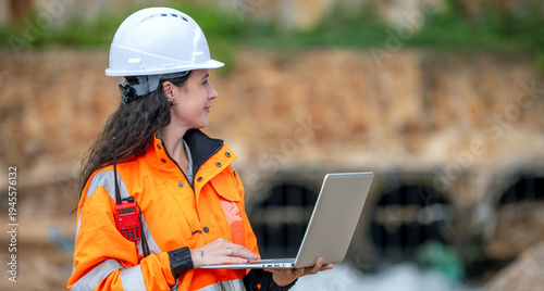 Female environmental scientist in safety gear standing and using laptop and radio for monitoring ecological impact inspection near industrial gas storage tank at fuel terminal site.