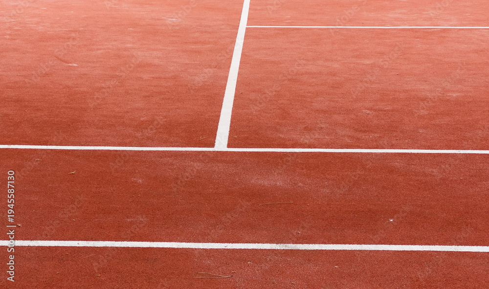 Fototapeta premium A clay court, marked with bright white lines, awaits the next set. Tennis balls scattered across the surface hint at a recent practice. Greenery surrounds the outdoor space.