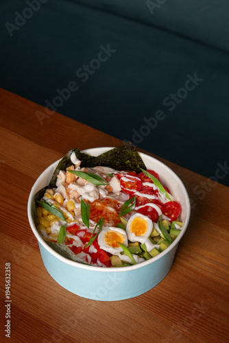 Fresh chicken poke bowl with egg, avocado, corn, cherry tomatoes, rice, nori and green onion served on wooden restaurant table