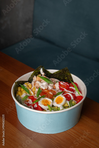 Fresh chicken poke bowl with egg, avocado, corn, cherry tomatoes, rice, nori and green onion served on wooden restaurant table