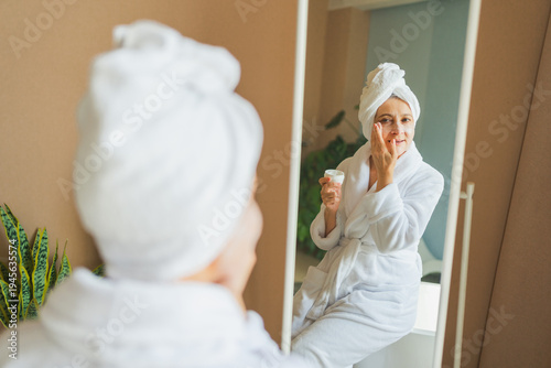 Mature adult old woman looking at mirror applying nourishing anti age face cream doing daily beauty routine. Senior woman with white mask creme on skin gets facial treatment Skin care aging skincare