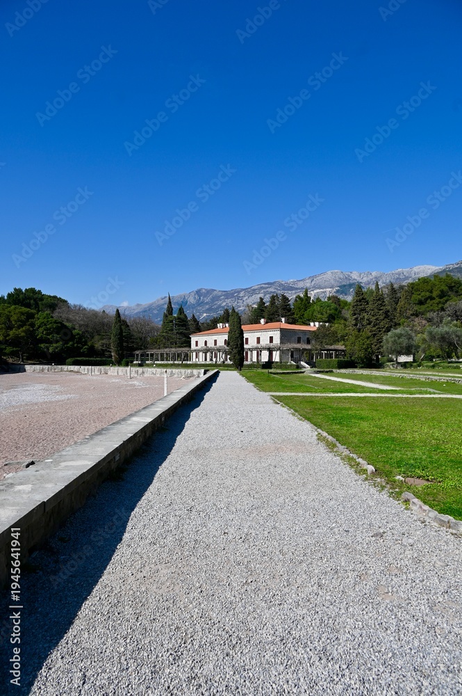 Obraz premium Wide angle view of a gravel path leading to the historic Villa Milocer building in a green park under a clear blue sky, Montenegro.