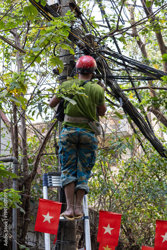 Electrician working on utility pole amid dense urban greenery: a worker in a red helmet and camouflage pants stands on a stepladder, repairing or inspecting a complex network of electrical cables