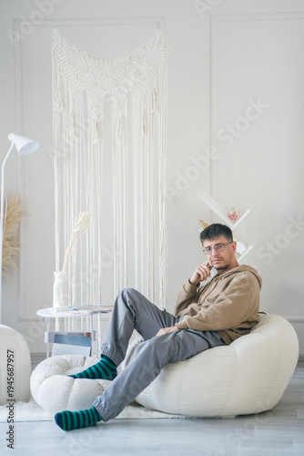 A relaxed man in a beige hoodie and glasses sits comfortably on a beanbag chair in a bright, minimalist room