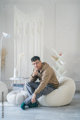 A relaxed man in a beige hoodie and glasses sits comfortably on a beanbag chair in a bright, minimalist room