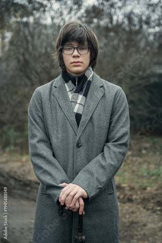 A stylish young man in a gray wool coat and scarf stands outdoors in autumn, holding a closed umbrella