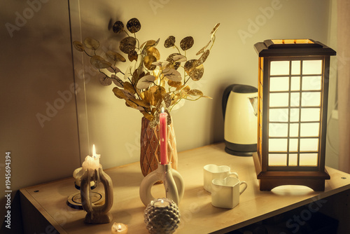A cozy evening scene featuring a wooden table with glowing candles, a decorative vase with gold foliage, heart-shaped mugs, and an elegant Japanese-style wooden lamp, creating a warm atmosphere.