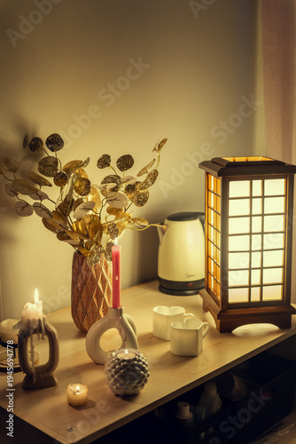A cozy evening scene featuring a wooden table with glowing candles, a decorative vase with gold foliage, heart-shaped mugs, and an elegant Japanese-style wooden lamp, creating a warm atmosphere.