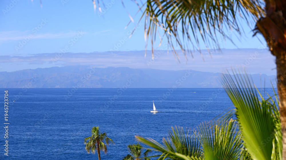 Obraz premium calm blue ocean stretching toward a distant horizon with a lone sailboat gliding across the water, framed by tall palm leaves and soft summer light that creates a peaceful and elegant seaside atmosphe