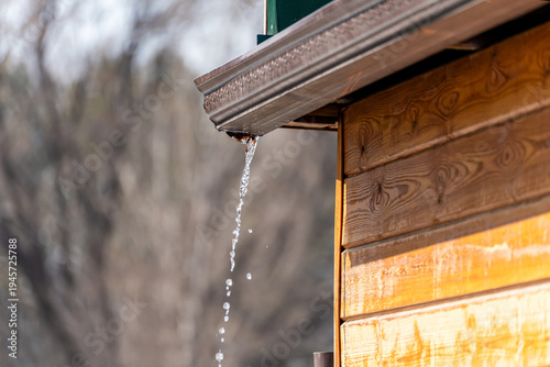 Close-up of water dripping from a brown metal rain gutter on a wooden house in bright sunlight. The image captures the natural process of thawing with a blurred natural background.