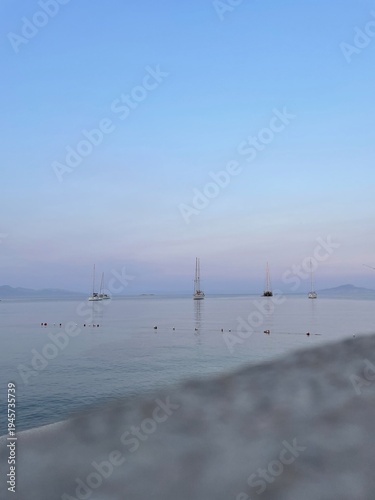 Sailboats anchored on calm water at pastel dawn horizon