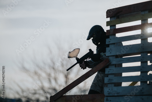 Paintball player wearing a helmet aims a marker from a wooden ramp. Outdoor action scene with a pallet barricade and light on the horizon.