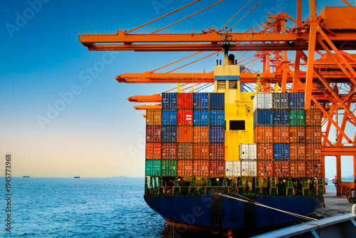 Rear view of a large container ship being discharged by massive orange quay cranes at a port terminal during sunset.
