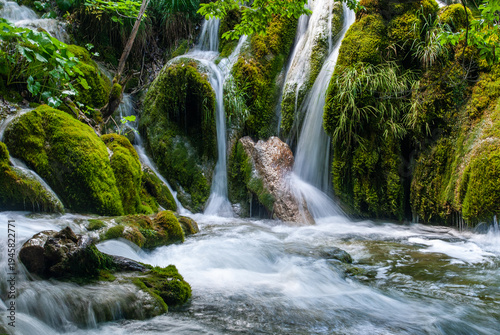 Serene forest lake with small waterfalls cascading over moss-covered rocks, surrounded by lush green vegetation and flowing gently through tranquil woodland. Plitvice Lakes National Park in Croatia.