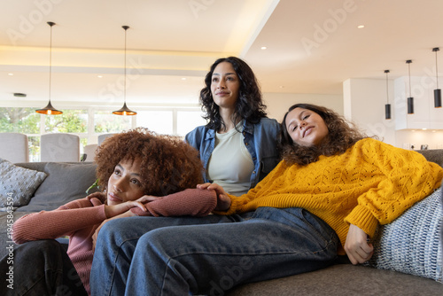 Diverse female friends sitting closely on gray sectional sofa, leaning and wearing mustard sweater
