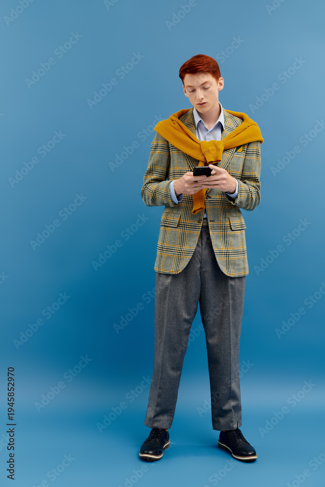 Naklejka premium Stylish young man checks smartphone while dressed in trendy attire against blue backdrop