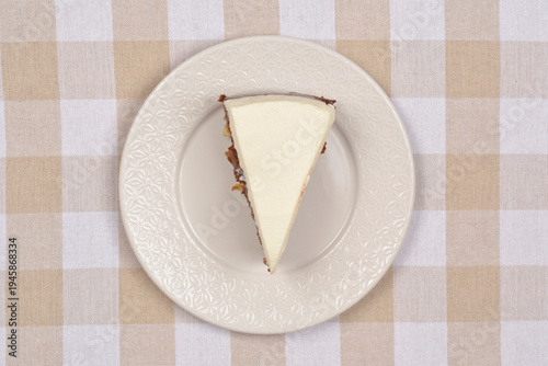 A top down view of a triangular slice of chocolate cream cake with white frosting served on a decorative plate over a beige and white checkered fabric background