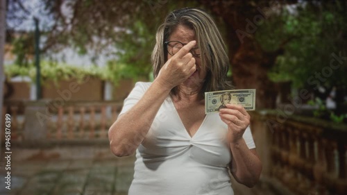 Hispanic woman with grey hair pinches nose while holding a twenty banknote in a building; money concern disgust.
