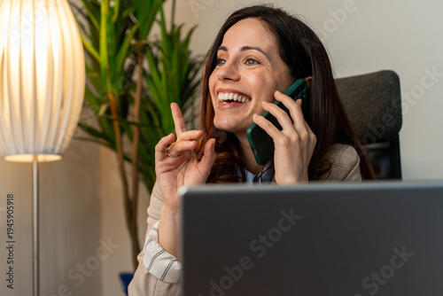 Happy smiling young business woman, 30s professional lady executive manager talking on the phone making business call on cellphone at work in office using laptop computer