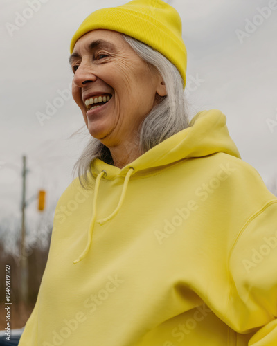 Elderly woman laughing outdoors in yellow beanie and hoodie