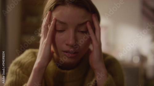 Handheld close up shot of distressed young adult woman holding head with hands experiencing intense anxiety while sitting in bedroom, subtle camera shake effect
