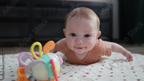 Baby during tummy time on a patterned mat, looking at a colorful rattle toy and drooling.