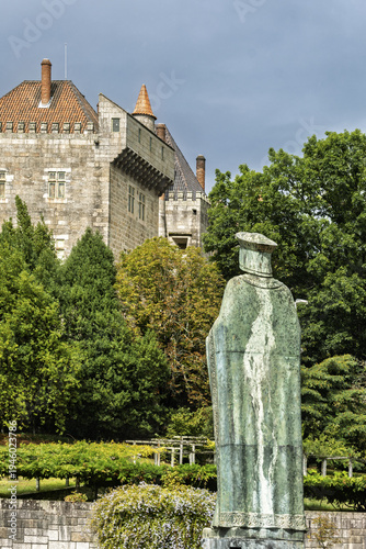 Guimaraes, Portugal, A hilltop stone castle rises above dense green tree, with a tall robed figure standing in the garden below, the cloudy sky giving the scene a calm historic weight.