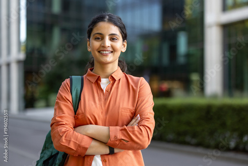 Young indian woman student standing outdoors with a backpack and crossed arms, confidently smiling at the camera, conveying themes of education, success, empowerment, and a bright future in the city