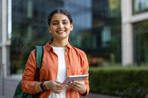 Young female student smiles holding a tablet and backpack on a modern university campus, enjoying outdoor learning, mobile education and confident, tech-savvy study life