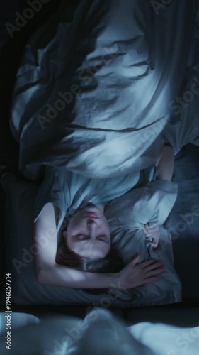 Vertical top view shot of young adult woman rolling from side to side on bed attempting to sleep then tossing pillow across bed nervously during night