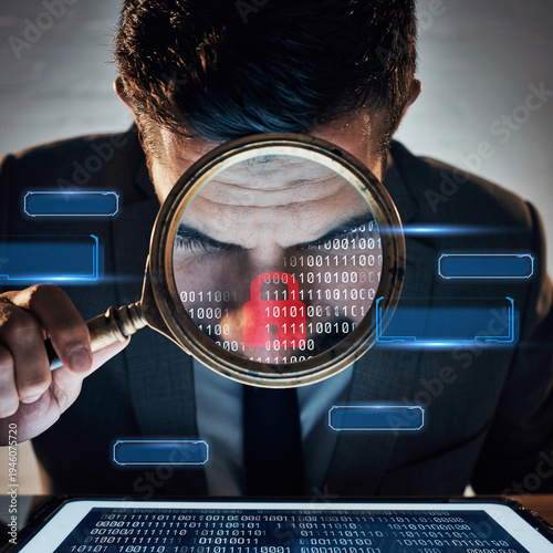 Close up of a man looking through a magnifying glass at binary code on a digital screen in a dark office setting