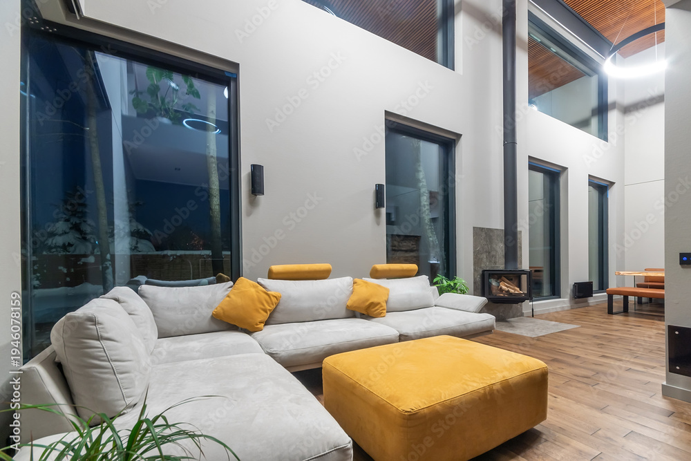 Naklejka premium Living room in a country house decorated in high-tech style. Modern architectural design, high ceiling, panoramic windows with a view of the evening winter landscape.
