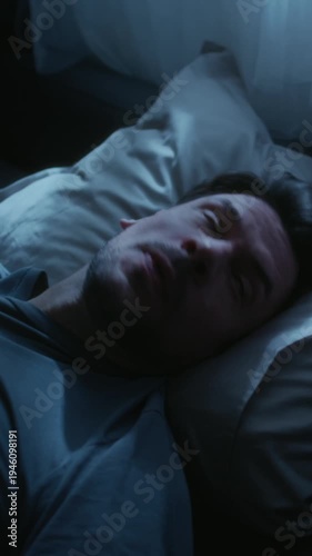 Vertical shot of adult man lying awake at night pressing hands to face in annoyance then turning over onto pillow trying to get comfortable and sleep during late night