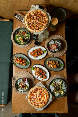 A wooden table is filled with different food items, including pizza, salads, and other dishes. People are enjoying a meal together in a casual dining space during lunch hours