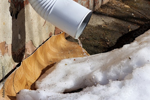 White downspout gushing water onto melting snow, signaling early spring thaw. Sunlight highlights wet surfaces and crumbling wall
