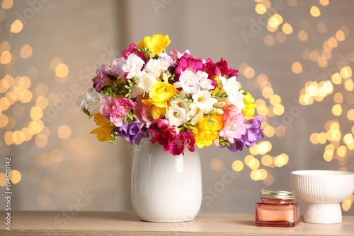Beautiful freesia flowers in vase and candle on wooden table indoors. Bokeh effect