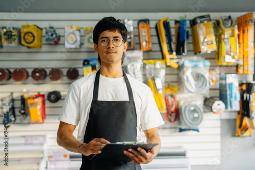 Fotografie Young man in apron holding clipboard, standing in front of shelves with products
