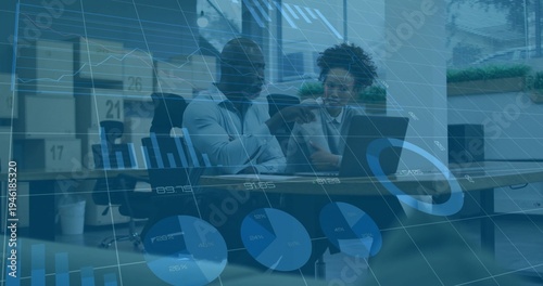 Pointing two colleagues in business attire reviewing laptop data at meeting table, with charts