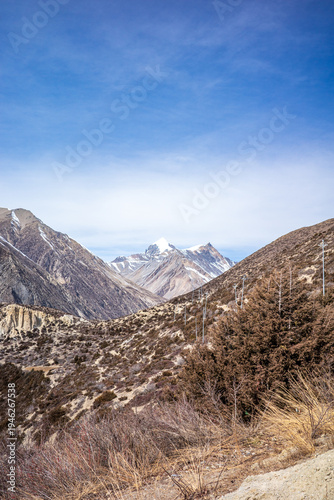 Scenic view of a dramatic Himalayan valley with snow-capped peaks rising in the distance in the Annapurna region of Nepal. Dry alpine hillsides and rugged mountain ridges frame the landscape