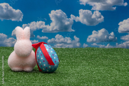 Pink fuzzy Easter bunny and decorated blue egg with red ribbon on green grass.