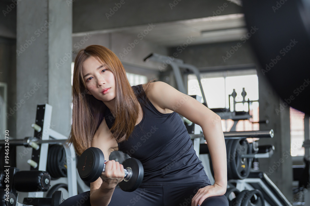 Fototapeta premium Asian athletic woman resting with dumbbell after intense workout session in modern gym, sitting on exercise bench while recovering during strength training routine.