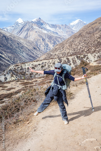 A joyful female hiker with a backpack and trekking poles celebrates on a mountain trail in the Annapurna region of Nepal. Surrounded by dramatic Himalayan peaks and rugged alpine landscapes
