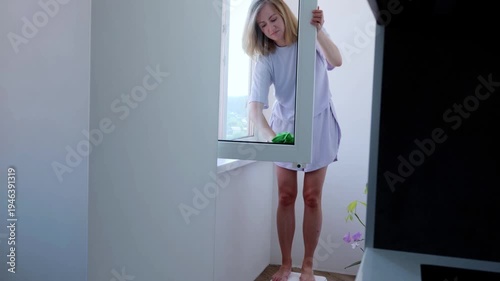 Woman wearing home clothes is cleaning windows with a sponge and detergent. She is standing barefoot on a small stepladder