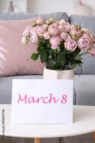 Vase with roses and card for International Women's Day on table in living room, closeup