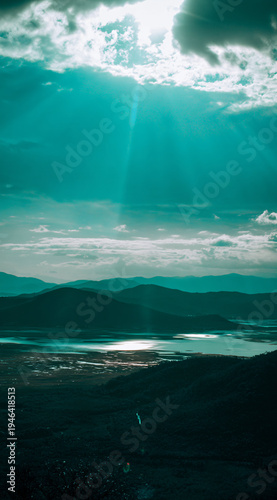 Stunning aerial view of a serene lake surrounded by mountains, with rays of sunlight breaking through clouds.